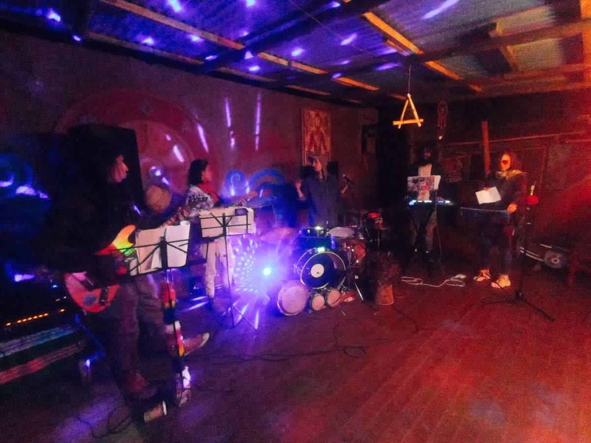 la noche en una sala de madera: en el escenario guitarristas con atriles, batería, teclado